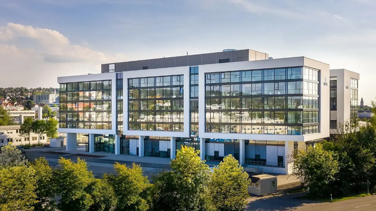Modernes, großes Bürogebäude mit viel Glasfassade, umgeben von Bäumen und Grünflächen, an einem sonnigen Tag.