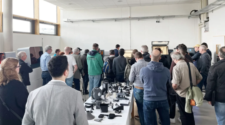 Eine Gruppe von Menschen in einem hellen Raum steht um einen langen Tisch mit verschiedenen technischen Geräten und Komponenten und sieht in eine Richtung, vermutlich einer Präsentation oder Vorführung folgend.