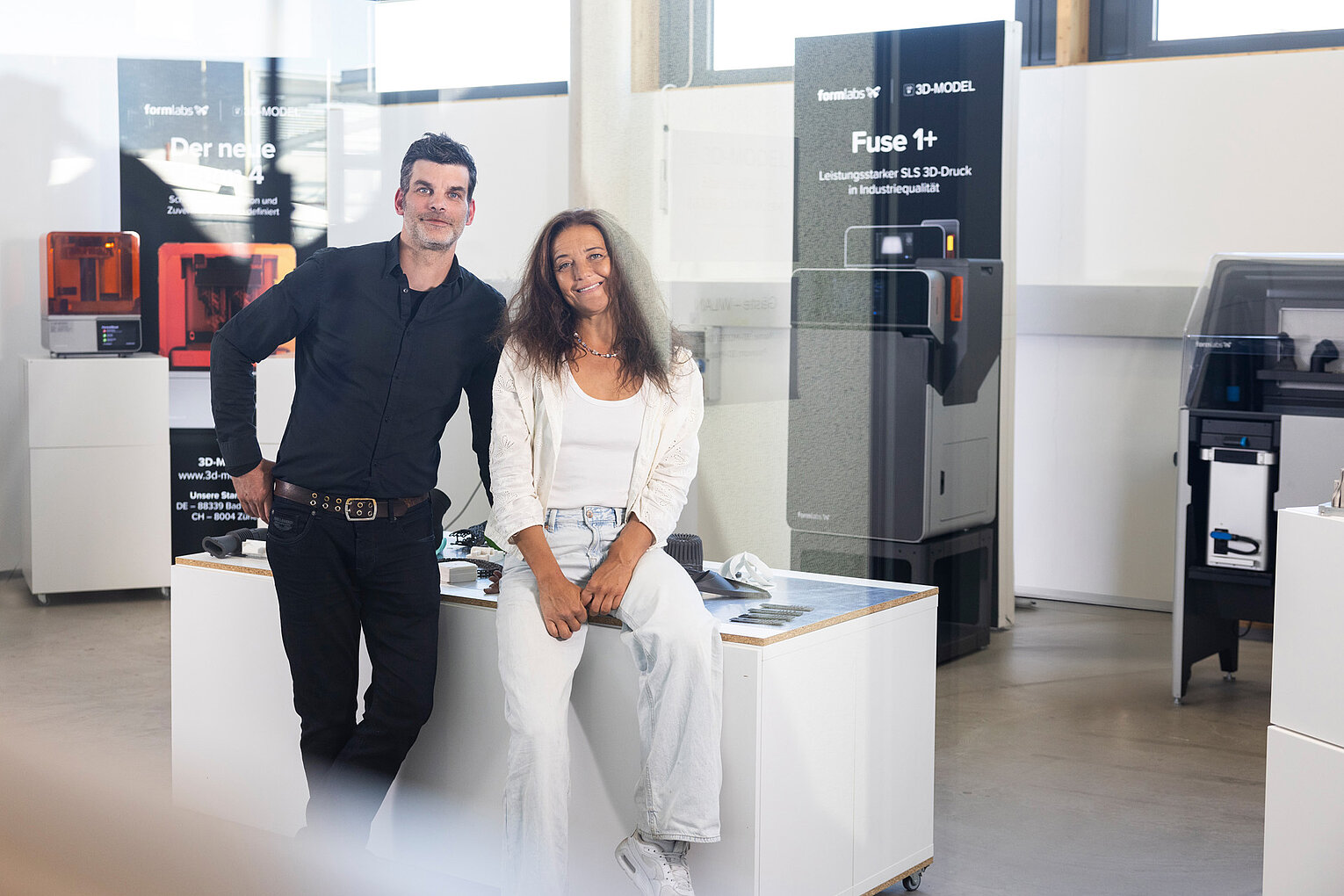 Zwei Personen lächeln in einem modernen Raum mit Geräten und Displays, die 3D-Drucktechnologie präsentieren.