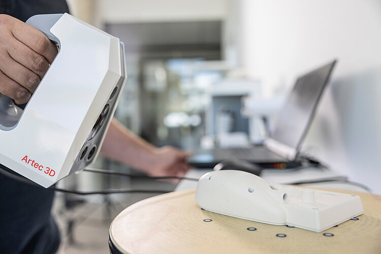 Person hält einen Artec 3D-Scanner und scannt ein weißes Modell auf einem Tisch in einem hellen Arbeitsraum mit einem Laptop im Hintergrund.