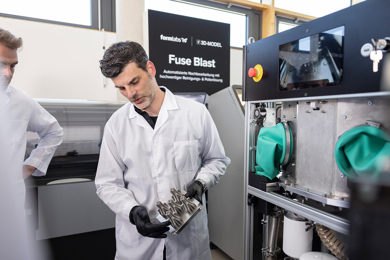 Ein Mann in Laborkittel und Handschuhen hält ein 3D-gedrucktes Metallteil neben einer industriellen Reinigungsmaschine in einem Labor oder einer Werkstatt.