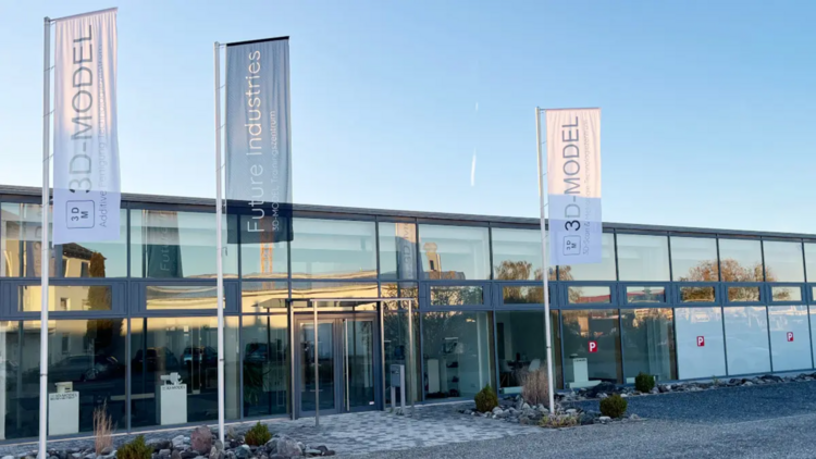 Gebäude mit großen Glasfronten und drei hohen Fahnen, zwei mit der Aufschrift 3D Model, eine mit Future Industries, unter blauem Himmel.