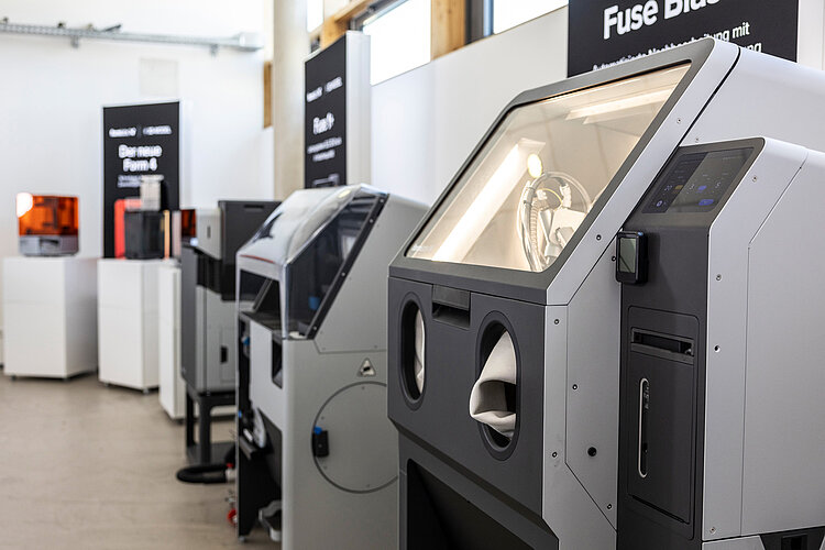 Mehrere 3D-Drucker unterschiedlicher Modelle stehen in einer gut beleuchteten Ausstellung oder Werkstatt nebeneinander.