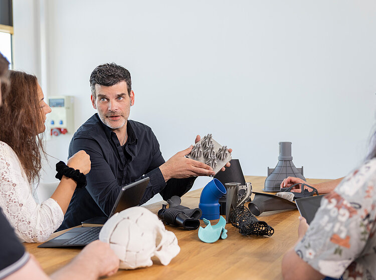 Eine Gruppe von Personen sitzt an einem Tisch und diskutiert 3D-gedruckte Objekte, während ein Mann ein Objekt zeigt und erklärt.
