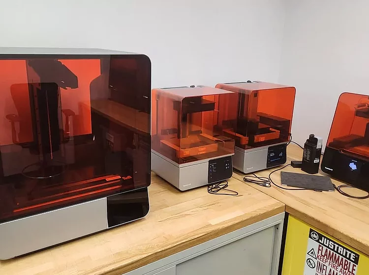 Mehrere 3D-Drucker mit orangefarbenen Schutzabdeckungen stehen auf einem Tisch in einem Labor oder Workshop.