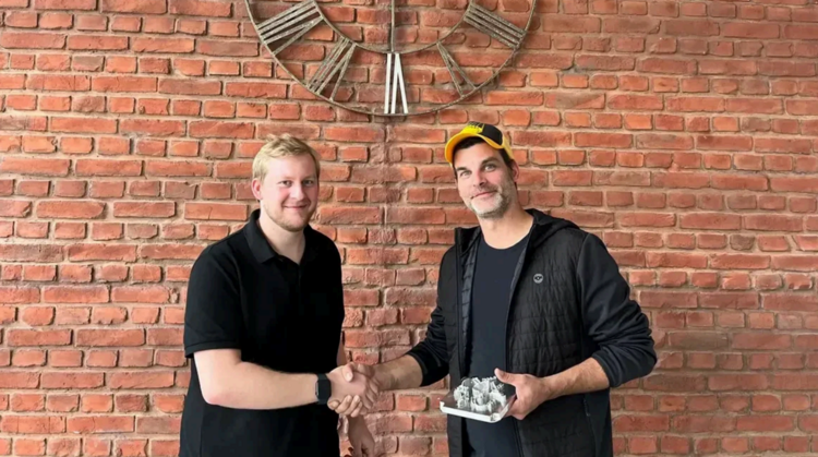 Zwei Männer stehen vor einer Backsteinwand und schütteln sich die Hand, einer hält eine kleine Skulptur.