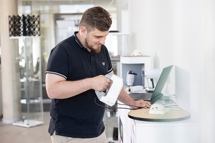 Ein Mann bedient ein elektronisches Messgerät, während er auf einen Laptop-Bildschirm schaut, in einem modernen Büro oder Labor.
