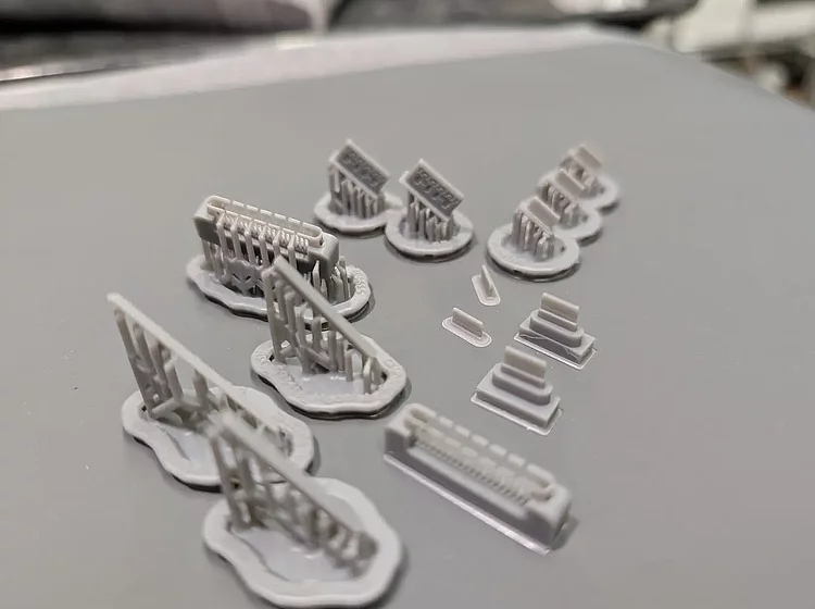 Mehrere kleine, graue 3D-gedruckte Objekte in verschiedenen Formen und Größen auf einer glatten Oberfläche angeordnet.