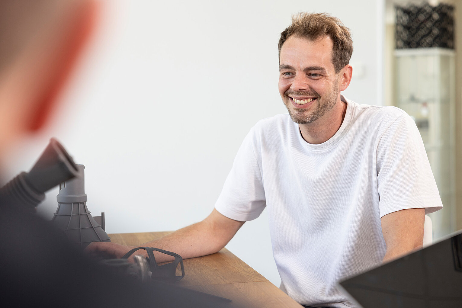 Ein lächelnder Mann in weißem T-Shirt sitzt an einem Tisch und unterhält sich mit einer anderen Person.
