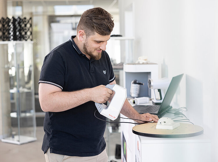 Ein Mann bedient ein elektronisches Messgerät, während er auf einen Laptop-Bildschirm schaut, in einem modernen Büro oder Labor.