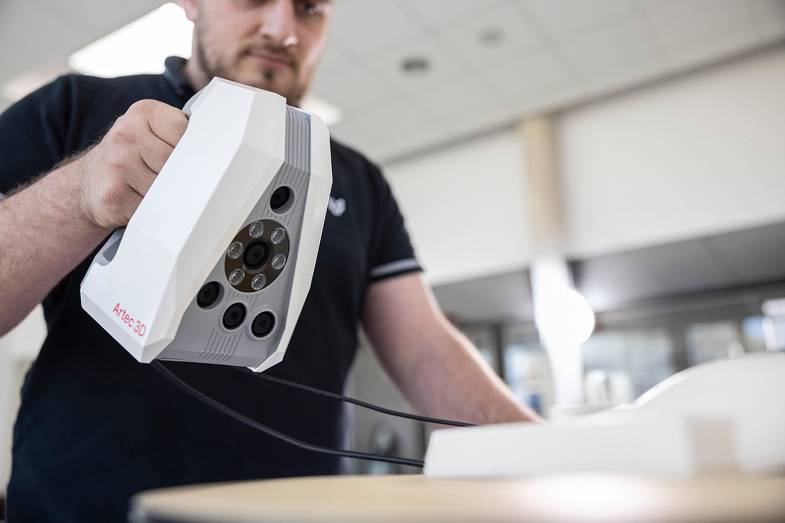 Eine Person hält ein weißes 3D-Scangerät, das auf einem Tisch über ein Objekt gerichtet ist.