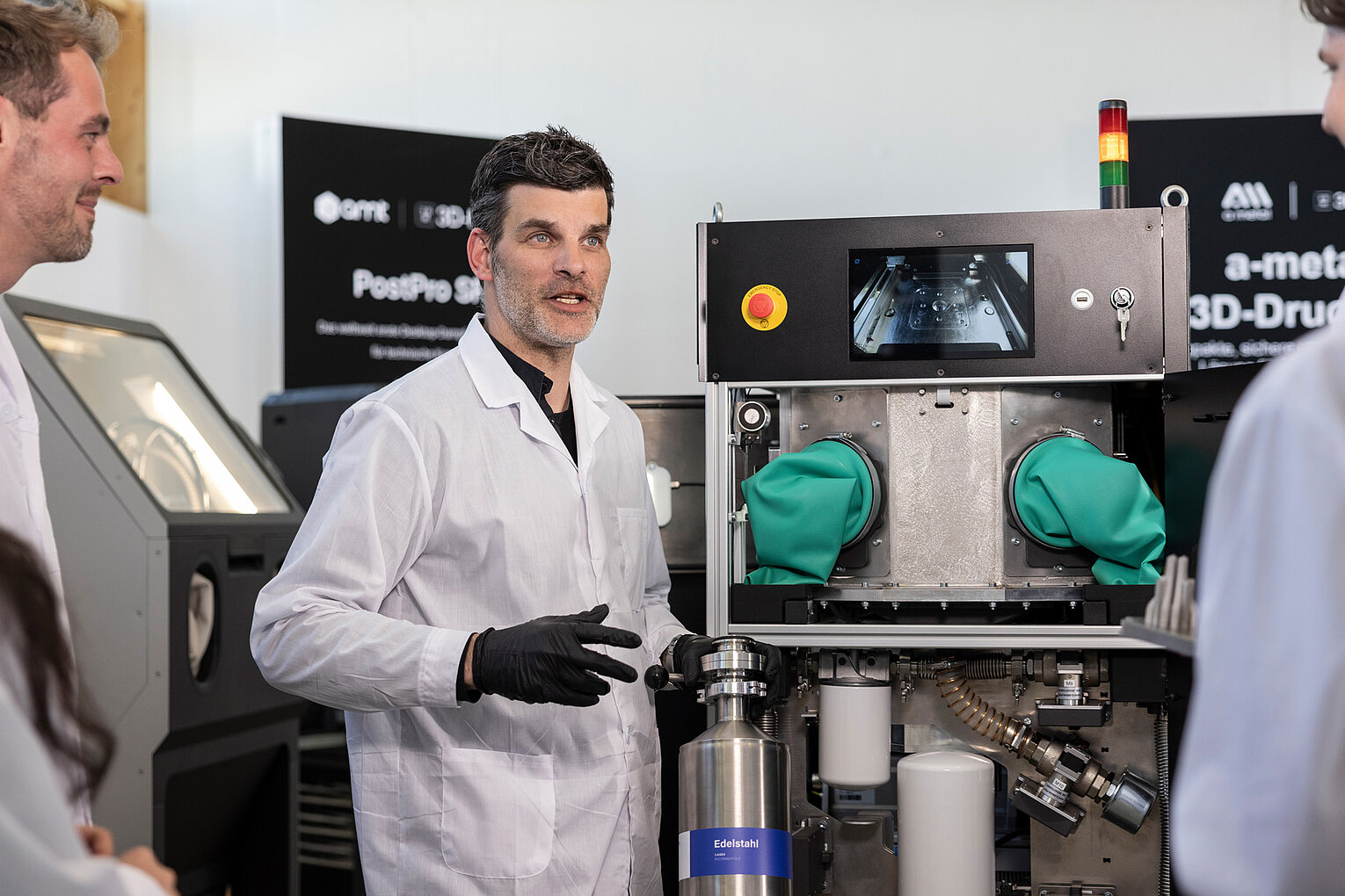Ein Mann im weißen Laborkittel erklärt zwei weiteren Personen die Funktionsweise einer industriellen Maschine mit grünem Handschuhfach und Display.