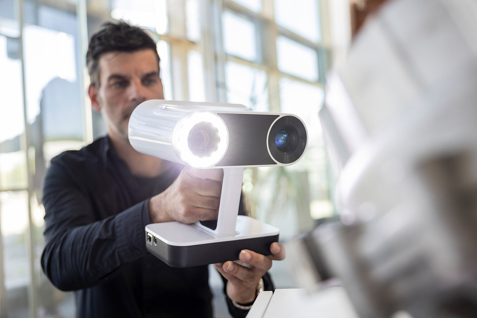 Mann hält ein weißes 3D-Scanner-Gerät mit Leuchtring und Kamera vor sich in einem hellen Raum mit Fenstern.