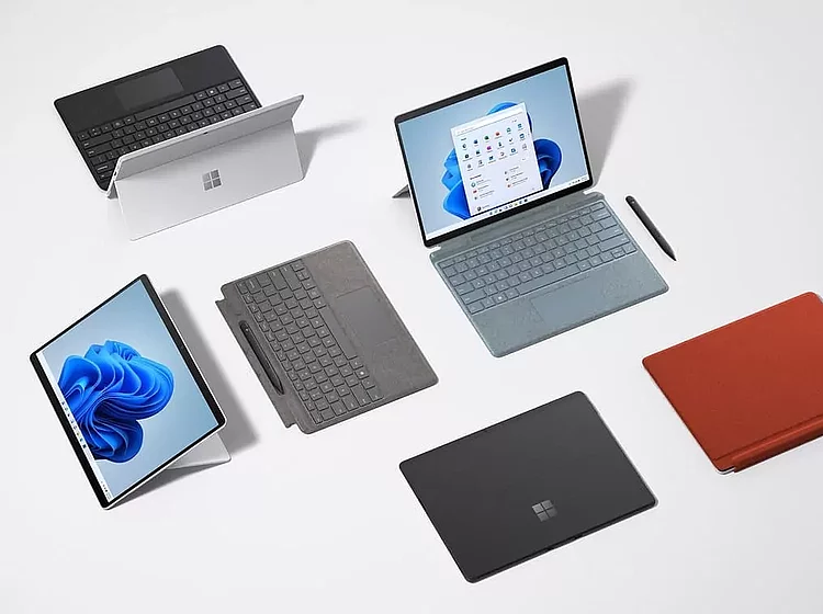 Verschiedene Microsoft Surface Geräte und Zubehörteile, darunter Laptops, Tablets, Tastaturen und Stylus, auf einem weißen Untergrund angeordnet.