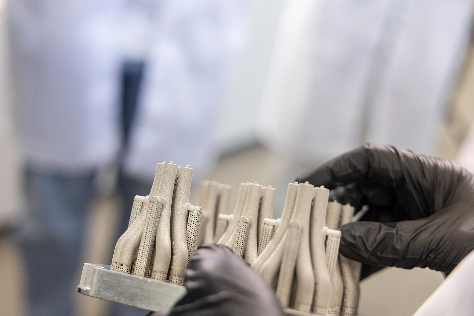 Hände mit schwarzen Handschuhen halten ein Bauteil mit mehreren 3D-gedruckten, röhrenförmigen Strukturen in einem Laborumfeld.