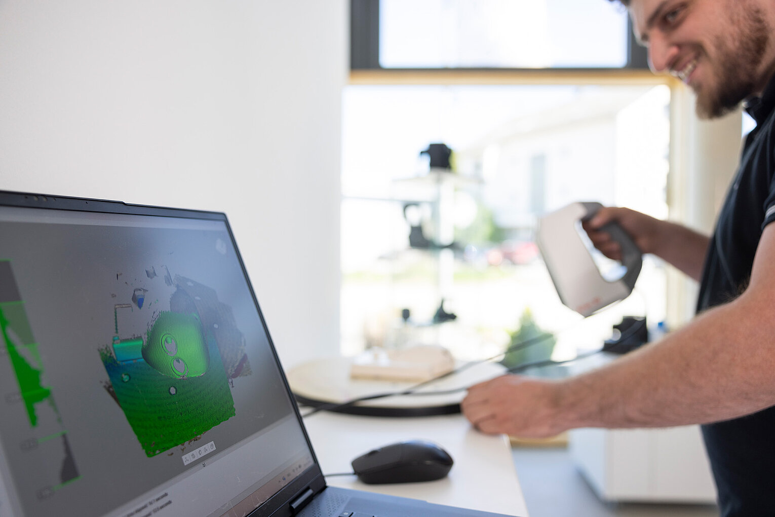 Mann mit Bart arbeitet am Computer und hält ein 3D-Scanner-Gerät in der Hand, während am Laptop ein 3D-Modell angezeigt wird.